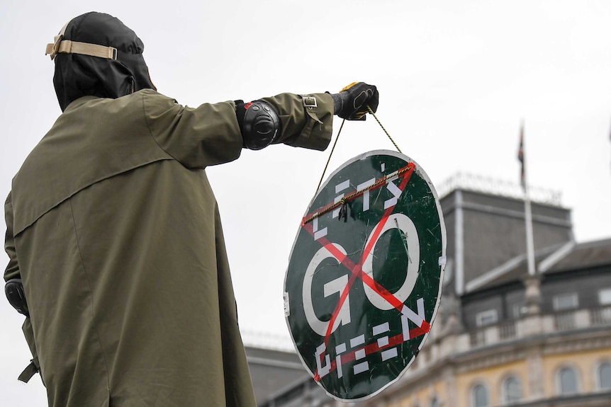 A man wearing a green trench coat and a gas mask over a black hood holds a black sign saying 'Let's Go Green'
