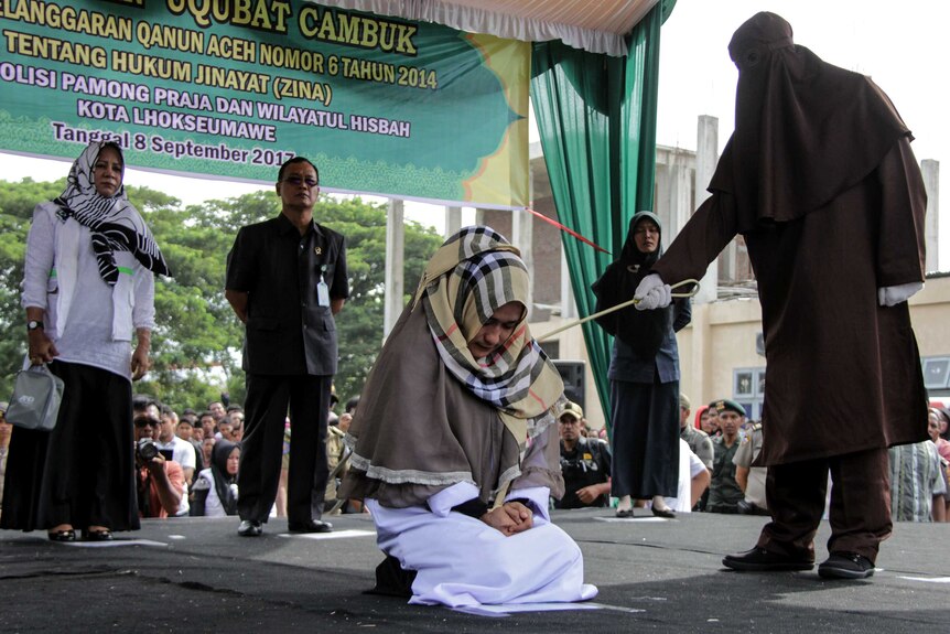 A kneeling woman winces as she is struck with a cane by a hooded man.