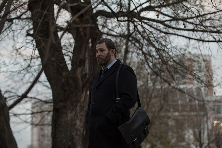 Still image from 2018 film Loveless of Aleksey Rozin looking forlorn and off camera in front of a tree and grey city skyline.