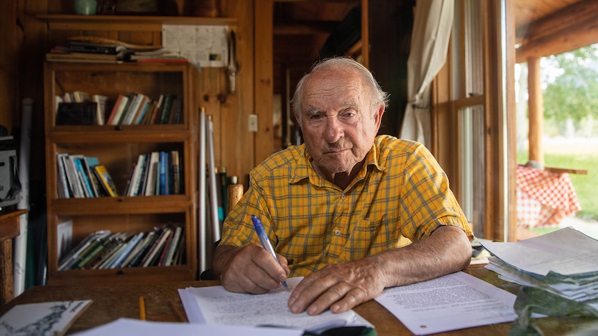 An elderly man in a yellow shirt sits at a wooden table writing a letter.