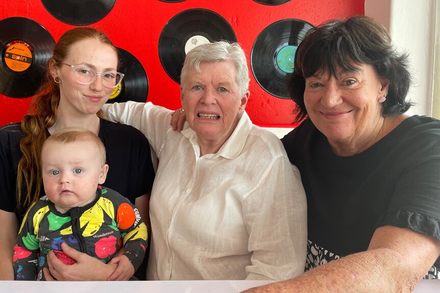 Three women and a baby sit in a diner in front of a red wall with records on it.