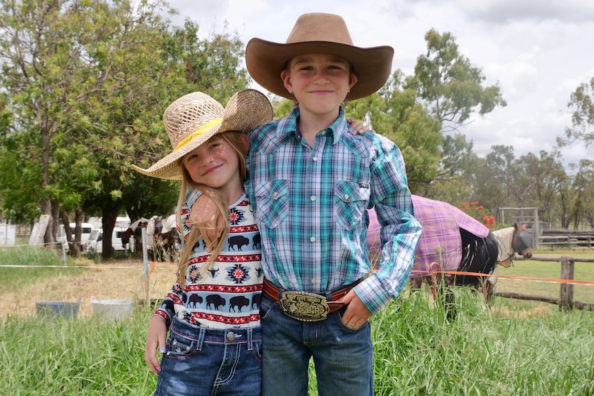 A boy and a girl stand in a field with their arms around each other.