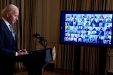 Joe Biden standing at a podium side-on with his head down, as members of his administration appear on a TV screen.