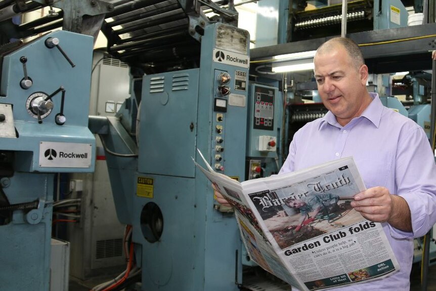 A man in a pastel purple business shirt holds a newspaper open while standing in front of printing press machinery