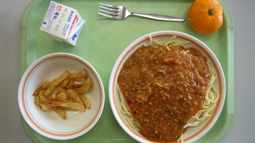 Spaghetti in a bowl on a green prison food tray, with an orange and a carton of milk.