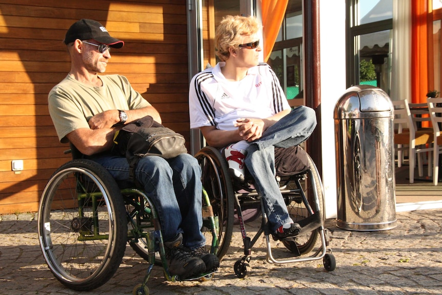 Two men in wheelchairs look to the right of the grame outside a house in Sweden.