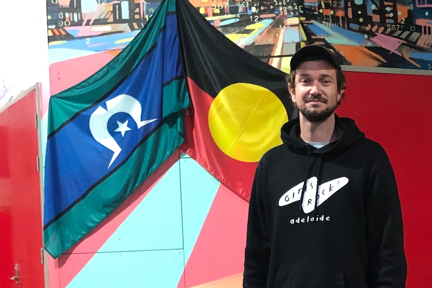 Nick O'Connor, in black jeans, hoodie, stands in a room covered in bright posters and Torres Strait Island and Aboriginal flags.