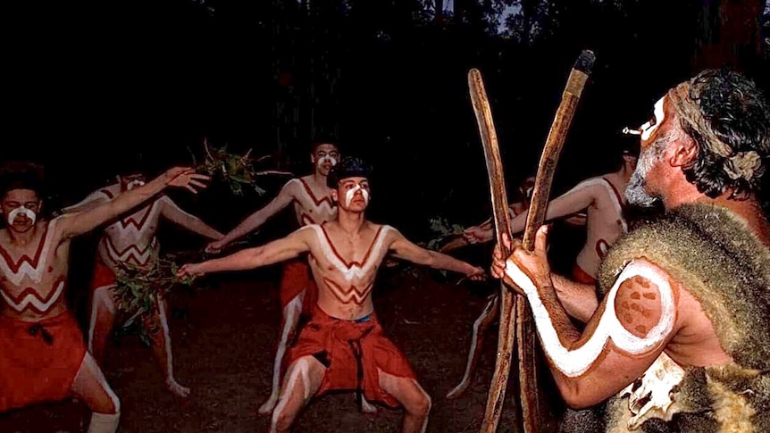 Indigenous dancers dance out bush at nighttime. The men wear body paint, dyed cloth and hold branches.