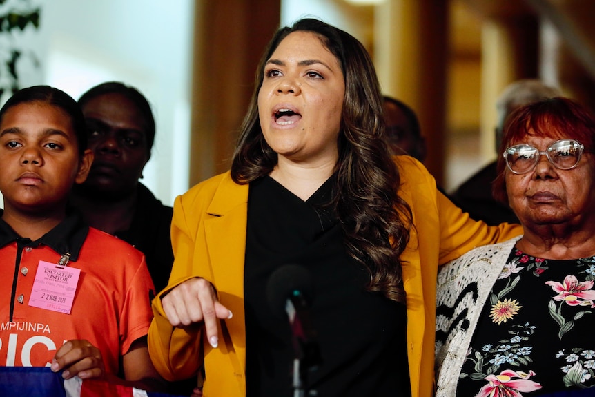 Indigenous woman Jacinta Nampijinpa Price speaks to a forum, with and Indegenous elder and child flanked either side.