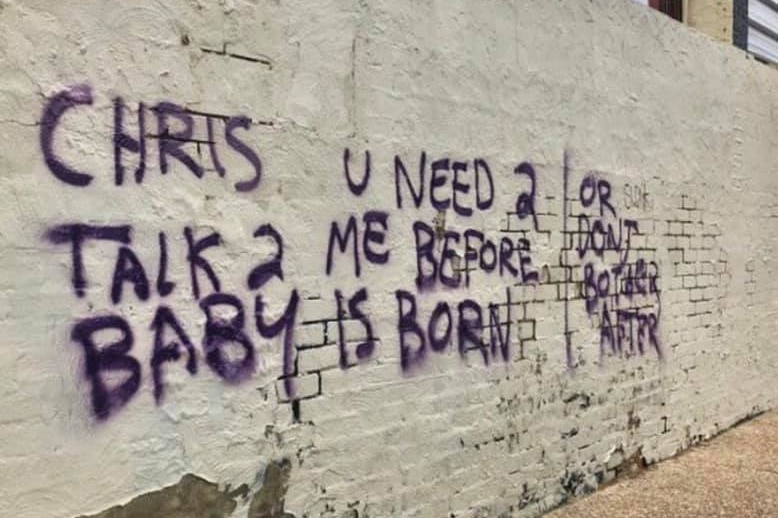 Graffiti sprayed onto a white wall reads 'Chris u need 2 talk 2 me b4 baby is born or dont bother after'.