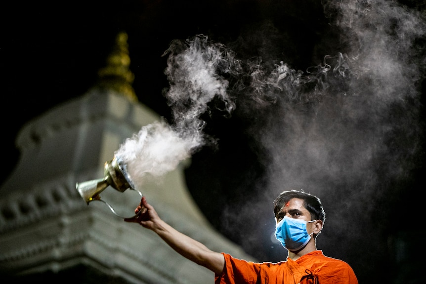 A man in a surgical mask and robes swings a smoking censer in front of a temple. 