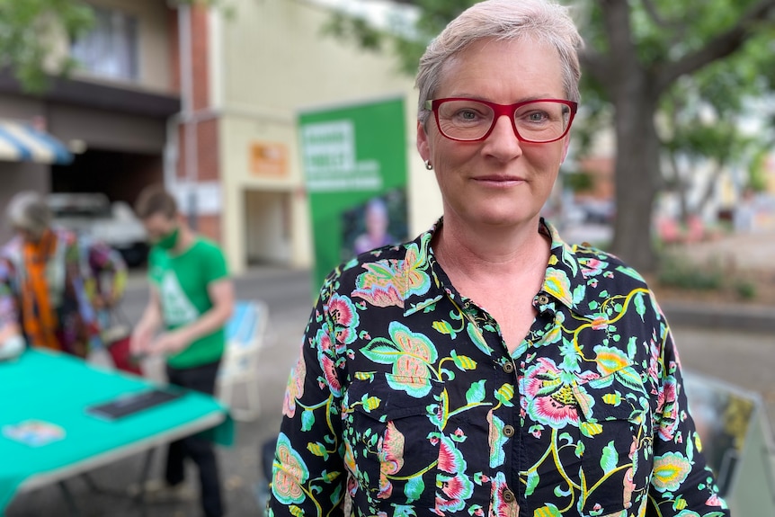 A woman with short, grey hair and stylish spectacles, wearing a brightly patterned shirt, stands on a street.