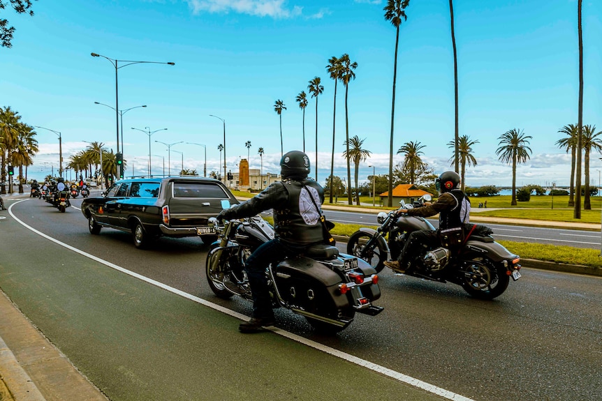 Two Aboriginal motorcyclists ride behind a black funeral car, past the palm-tree lined esplanade in St Kilda.