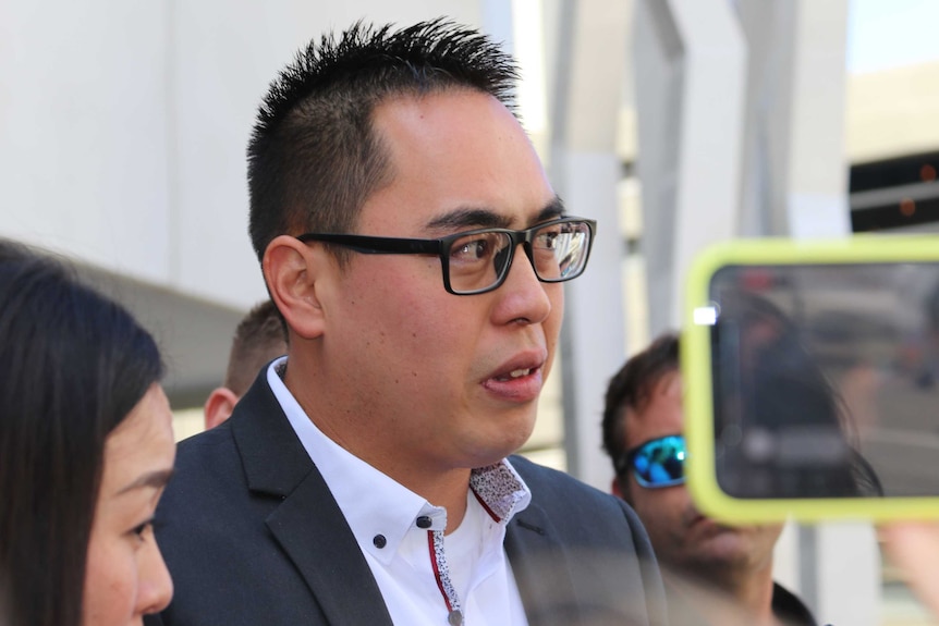 A tight head and shoulders shot of Andrew Doan wearing a suit and glasses speaking to media outside court.