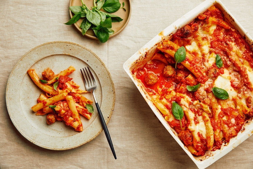 A serving of pasta al forno on a plate topped with basil leaves, from a dish of baked pasta with sausage meatballs.