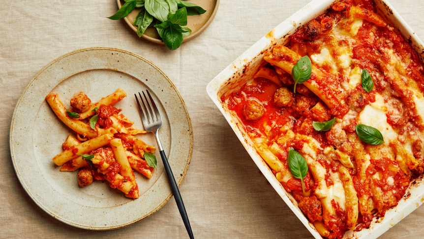 A serving of pasta al forno on a plate topped with basil leaves, from a dish of baked pasta with sausage meatballs.