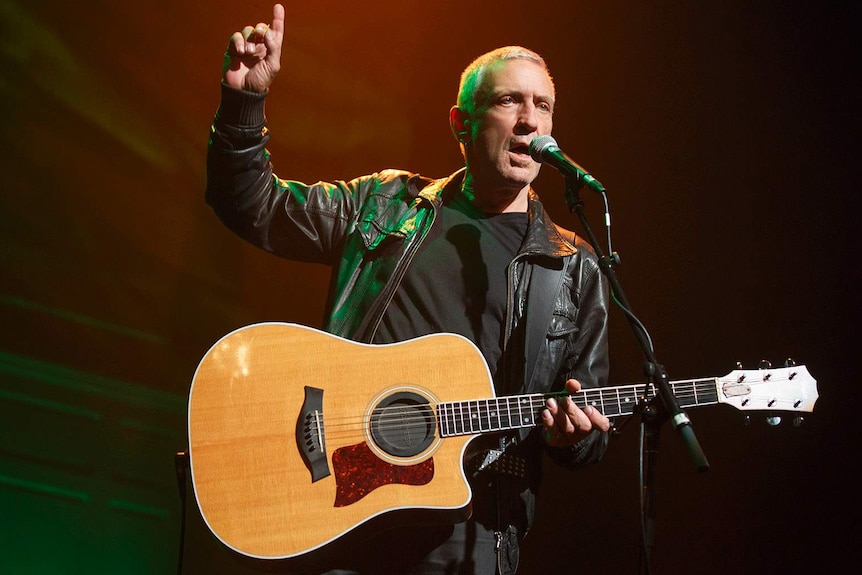 An actor holding his arm in the air while playing a guitar and singing