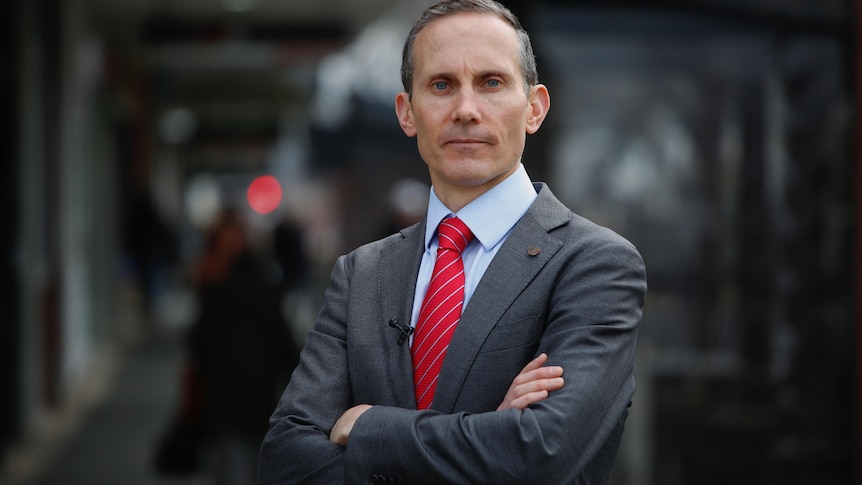 Andrew Leigh wearing a dark grey suit, blue shirt and red tie, standing with arms folded.