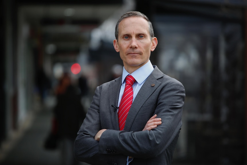 Andrew Leigh portant un costume gris foncé, une chemise bleue et une cravate rouge, debout, les bras croisés.