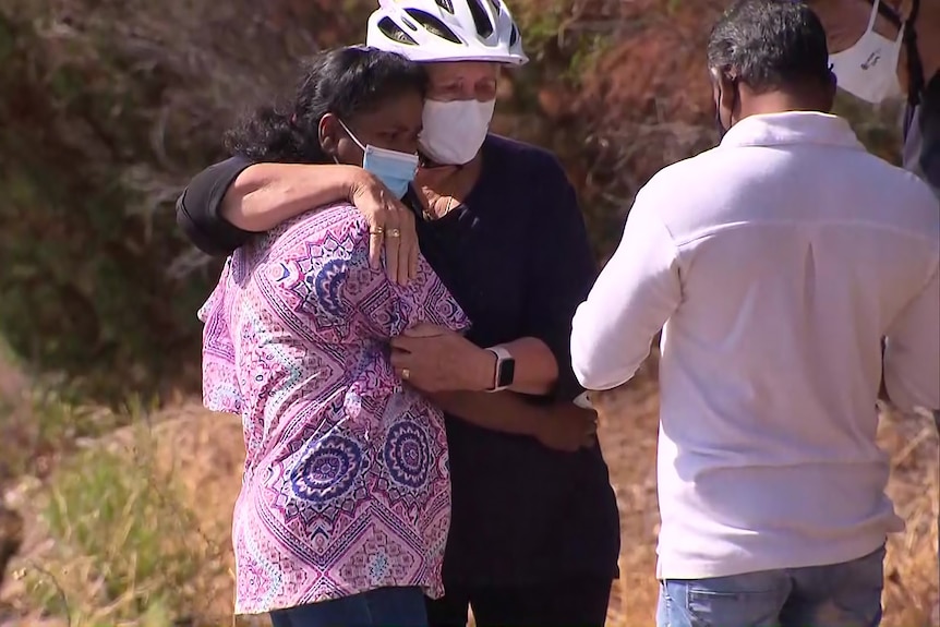 Two women, one of them wearing a bike helmet, hugging outdoors.