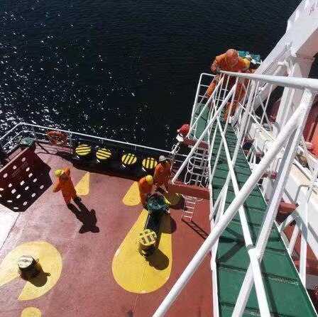 From a high vantage point atop a ladder, you look down at seafarers in orange boiler suits on a red ship's deck.