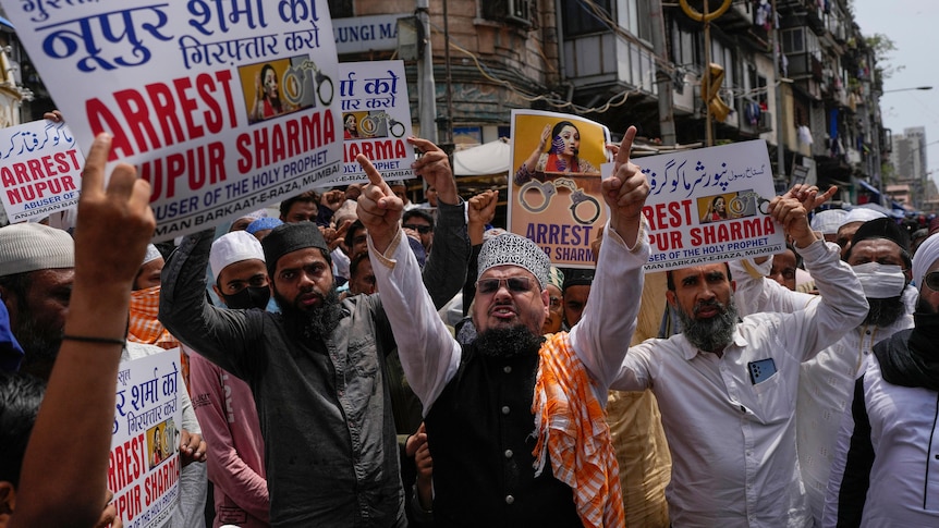 Bearded men in Islamic garb stand on a street holding multilingual placards calling for the arrest of Nupur Sharma