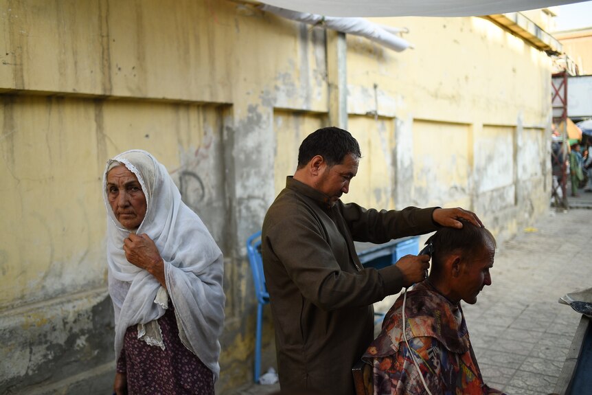 A woman walks by holding her white hijab as a man cuts the hair of a man sitting in front of him.