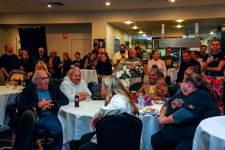 A group of people sit in an indoor function room, talking and laughing.