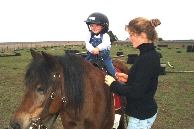 young girl rides a pony in a grassy field with her mother who wears a black turtleneck
