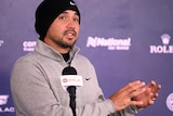 Wearing a beanie, Jason Day gestures with his hands while speaking in front of a microphone