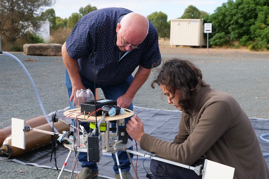 An older man bends down to look at a wooden and aluminium contraption with metal legs being built by his son.