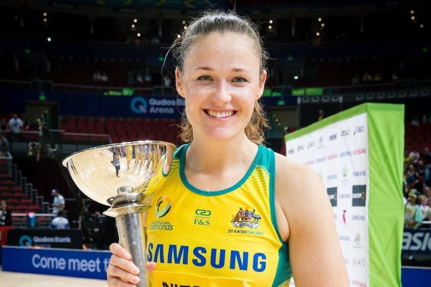 A woman wearing the Australian netball uniform smiles as she holds a trophy.