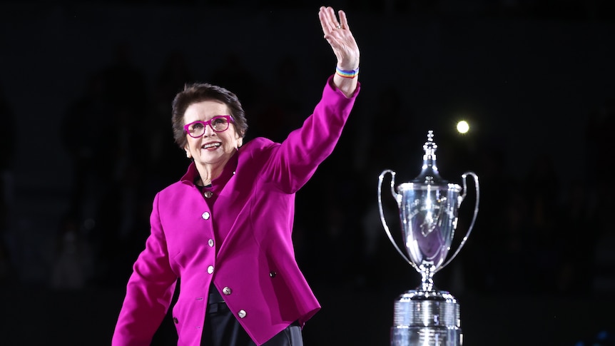 A former champion tennis player wearing pink glasses and a pink jacket waves as she stands with a big trophy.in the bqackground.