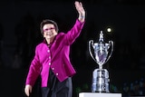 A former champion tennis player wearing pink glasses and a pink jacket waves as she stands with a big trophy.in the bqackground.