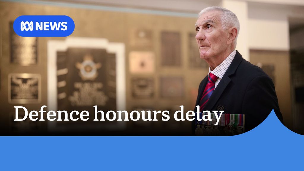 Defence honours delay. Image of an elderly man wearing a suit standing in front of an awards cabinet. 