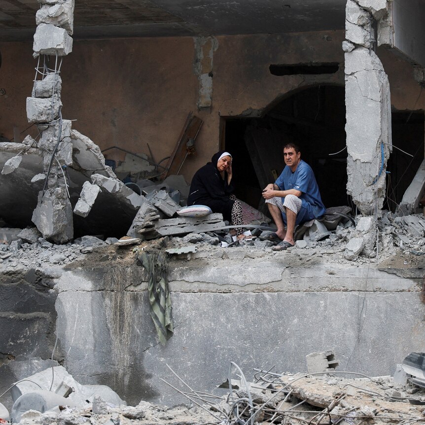 Two Palestinians - a man and a woman - sit among the rubble of a damaged residential building,