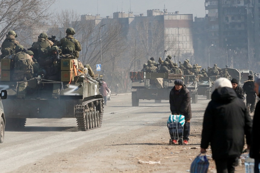 A convoy of tanks marked with white 'z's drive past a group of people talking along a dusty road.