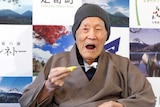 Masazo Nonaka sits in a wheelchair with a brown coat and teal blanket while eating a teaspoon of a cake sitting in front of him.