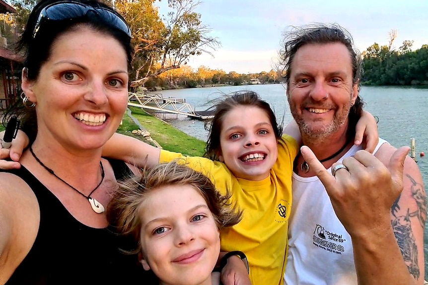 A man and woman with their children in front of the Murray River. The man is throwing a shakas and they are all smiling.