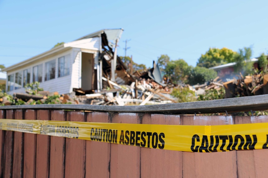 Blurred partly demolished weatherboard home in background, with yellow tape on fence in foreground, saying 'caution asbestos'.