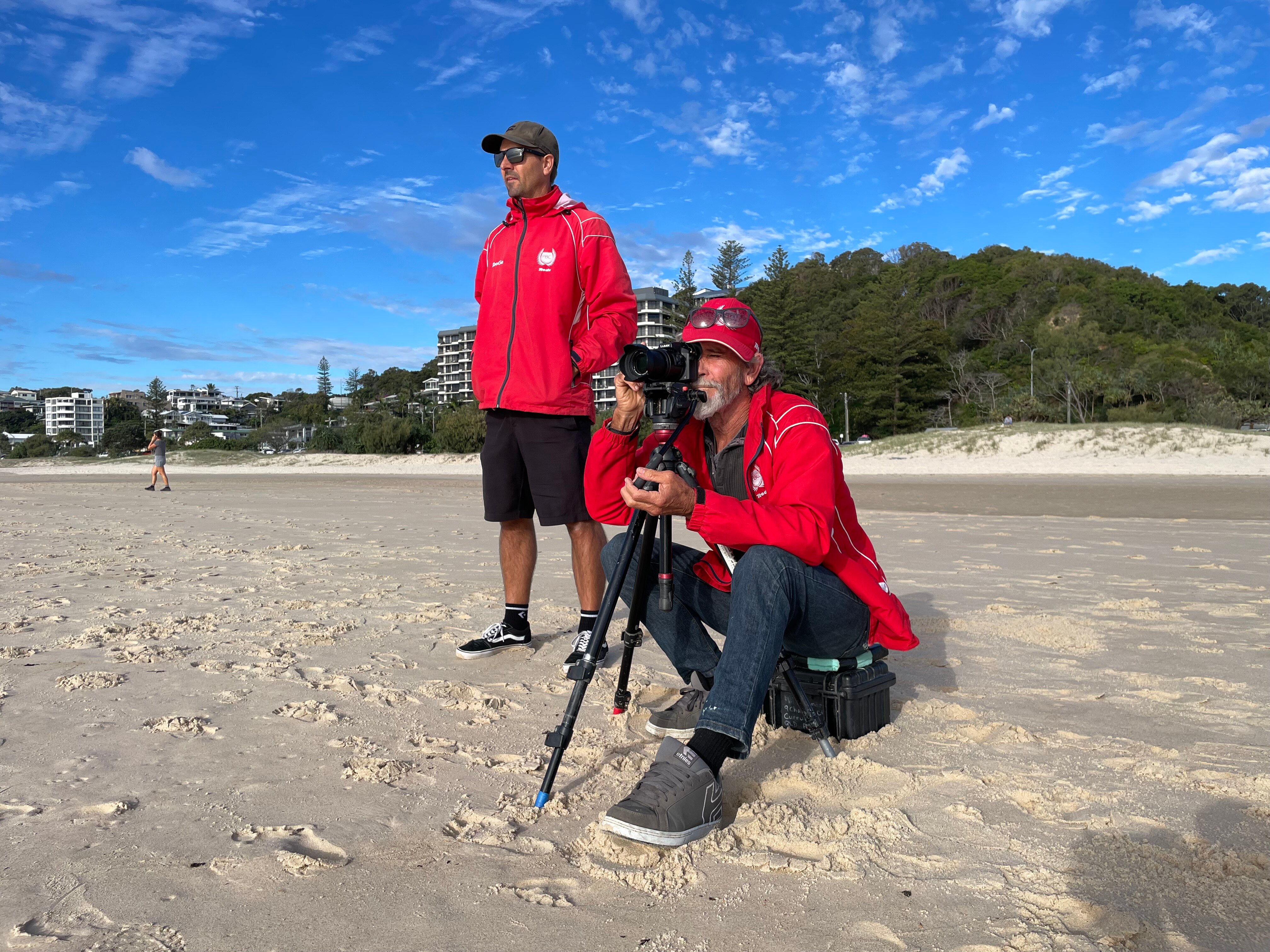 two school surf coaches on the beach one looking down the lense of a camera towards the waves