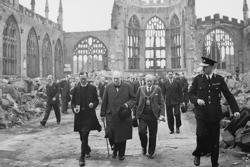 A black and white image showing Churchill walking in a group through the ruins of a cathedral.