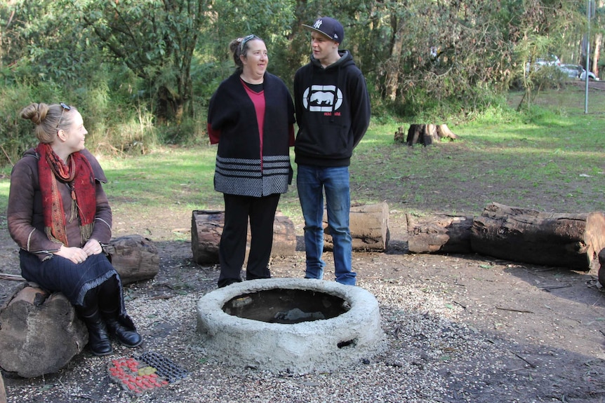 Three people stand around a campfire, smiling and talking.