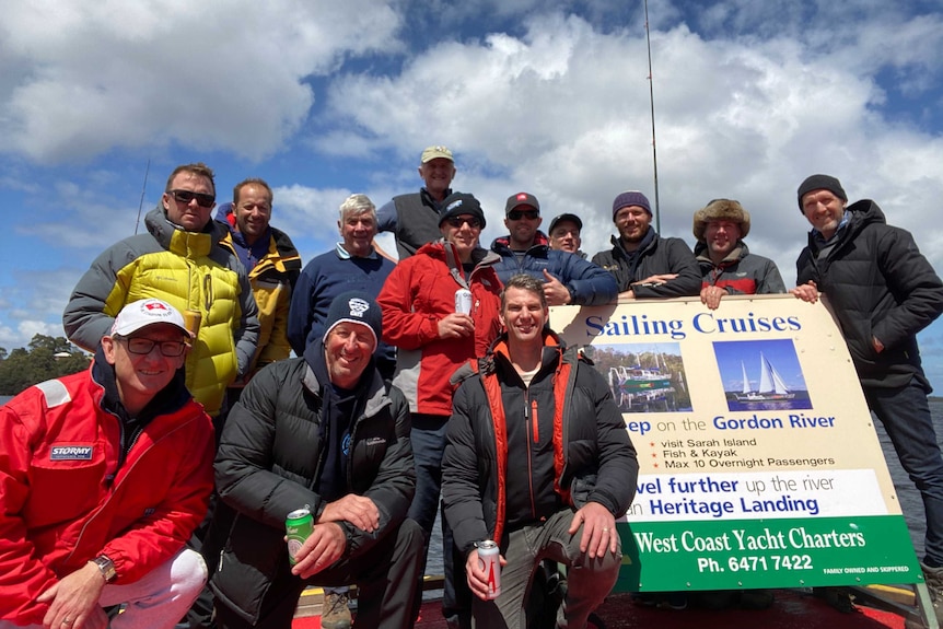A group of men in outdoor clothing smile as they sit around a sign for Gordon River sailing cruises