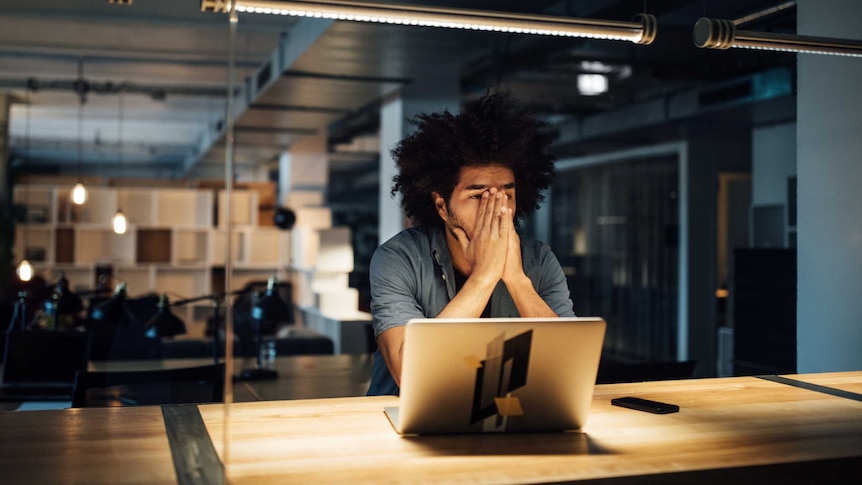 In a dark office, a man with curly black hair puts his hands over most of his face. A laptop sits in front of him.