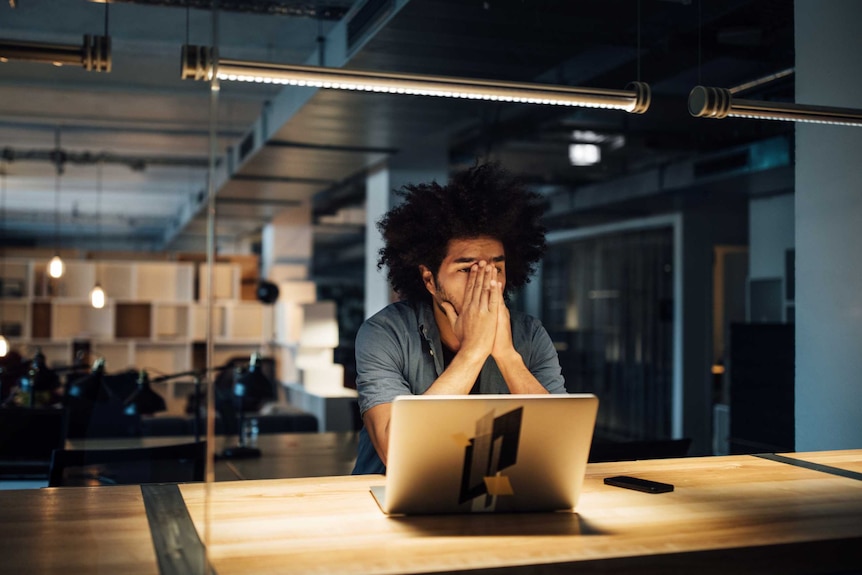 In a dark office, a man with curly black hair puts his hands over most of his face. A laptop sits in front of him.