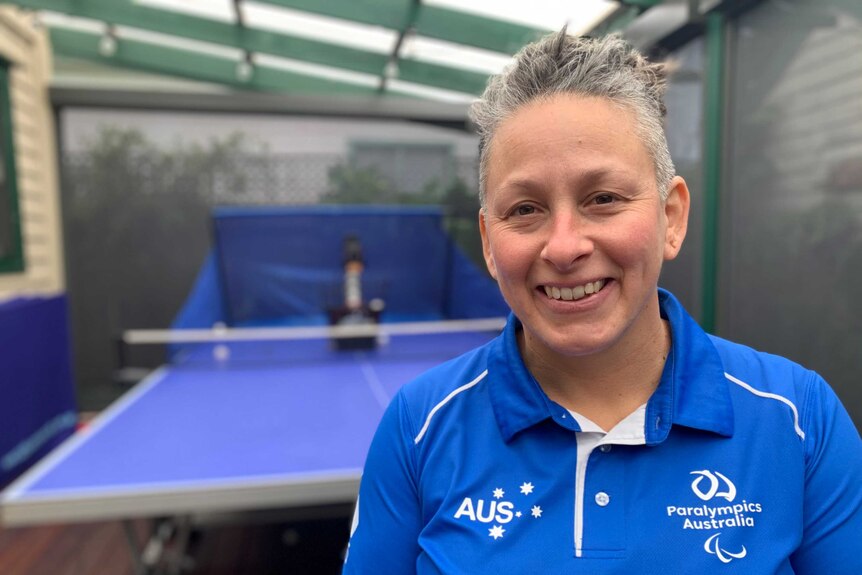 a woman smiles into the camera. a table tennis table is behind her.