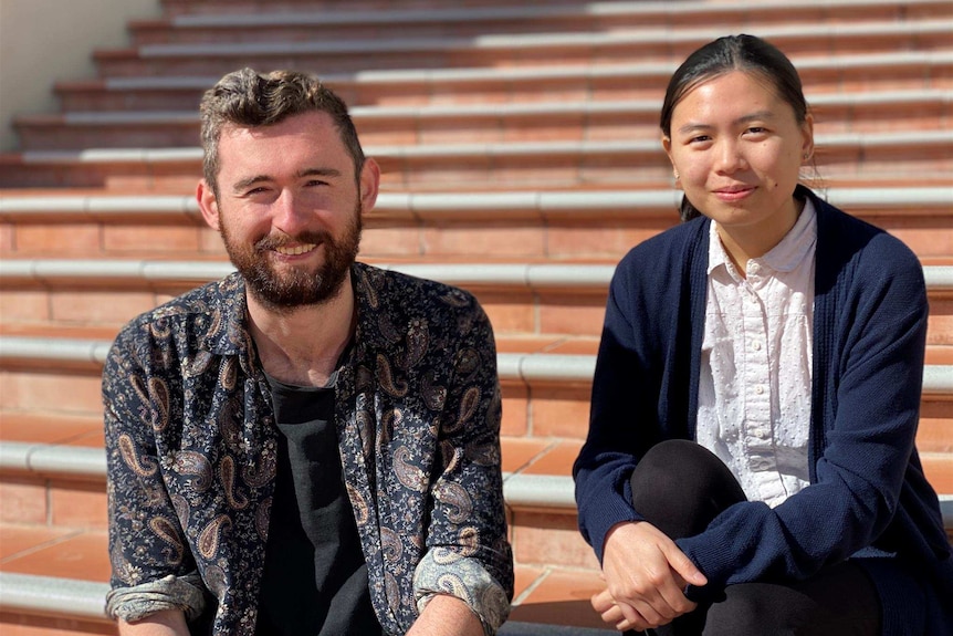 A white man sits next to an Asian woman on a set of stairs. They both look to the camera and smile, sunshine is on their faces.