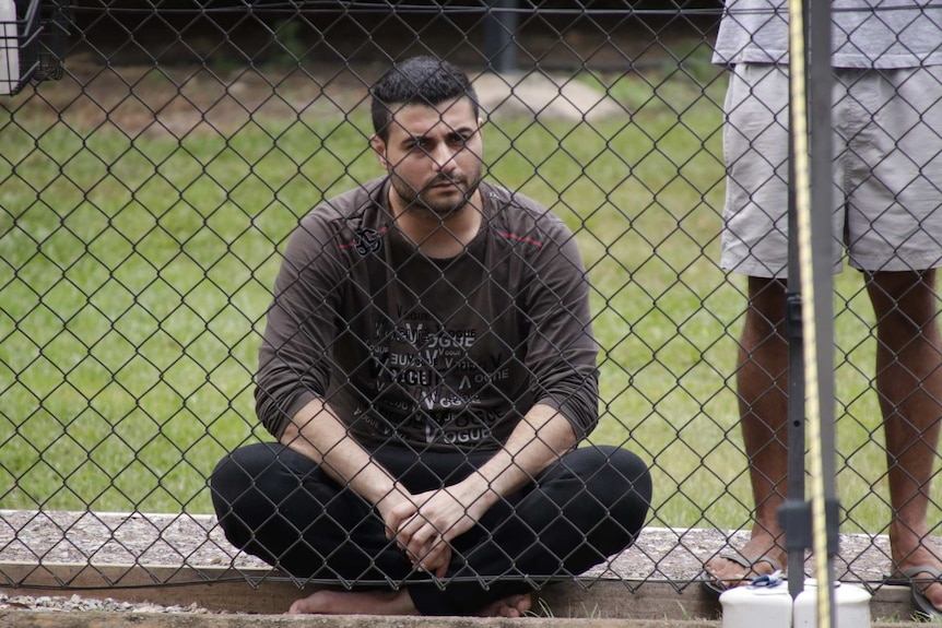 A man wearing a brown shirt, black jeans and no shoes sits on the ground with his legs crossed, behind a high wire fence.