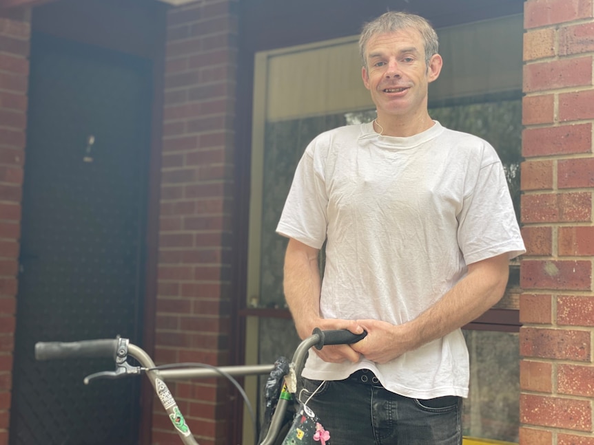 A man in a white shirt stands in front of a red brick unit holding his bike and smiling